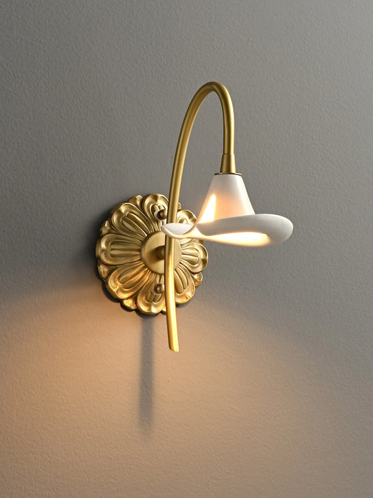 A gold floral wall sconce with a white ceramic shade and curved arm, mounted on a grey wall, emitting warm light.
