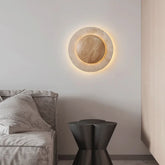 A modern round wall sconce made of natural stone with a wooden accent, illuminated by a warm LED halo effect, mounted on a white wall above a grey sofa.