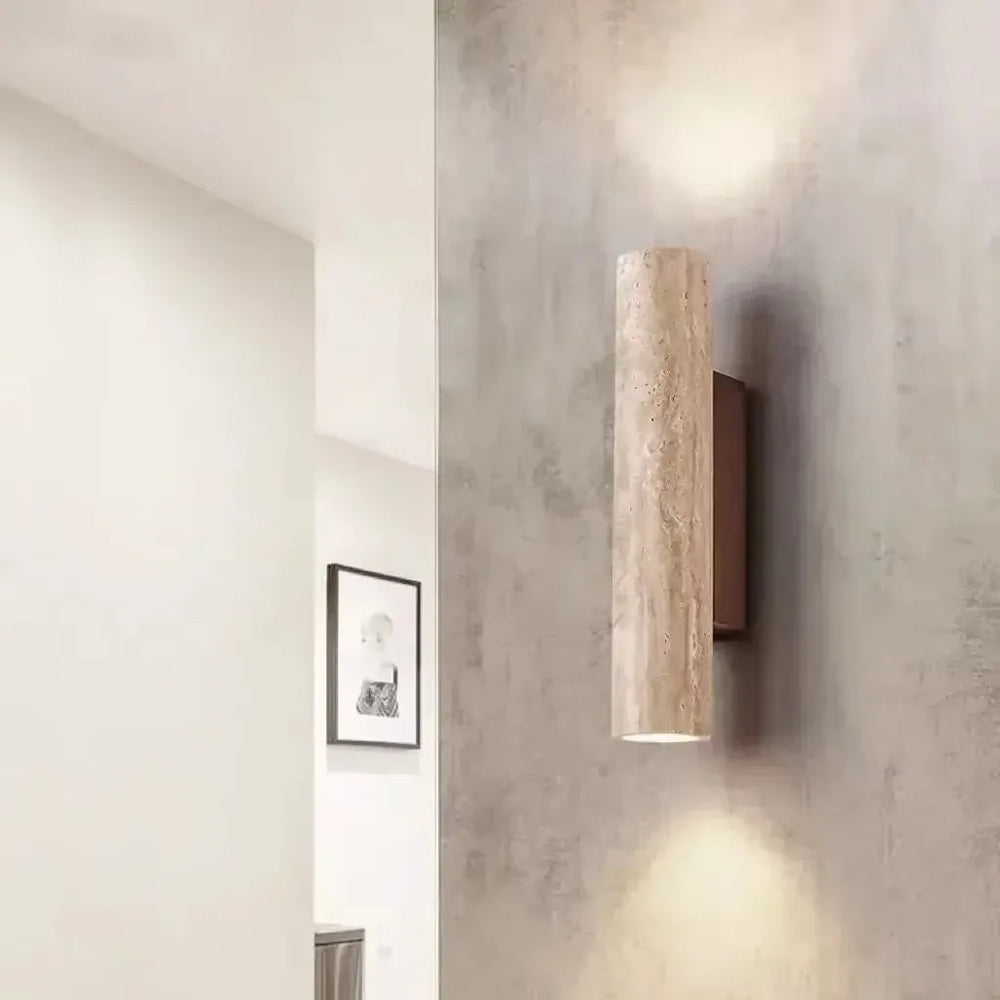 A cylindrical stone wall sconce mounted on a textured gray wall, casting warm light upwards and downwards in a hallway or entryway.