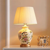 A ceramic table lamp with a white pleated fabric shade and a hand-painted floral and bird design on its base, placed on a white table.