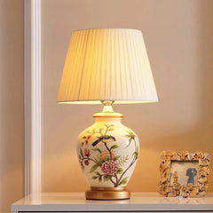 A ceramic table lamp with a white pleated fabric shade and a hand-painted floral and bird design on its base, placed on a white table.