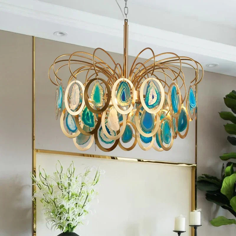 A gold chandelier adorned with blue and green agate slices hangs in a well-lit room with a large plant.