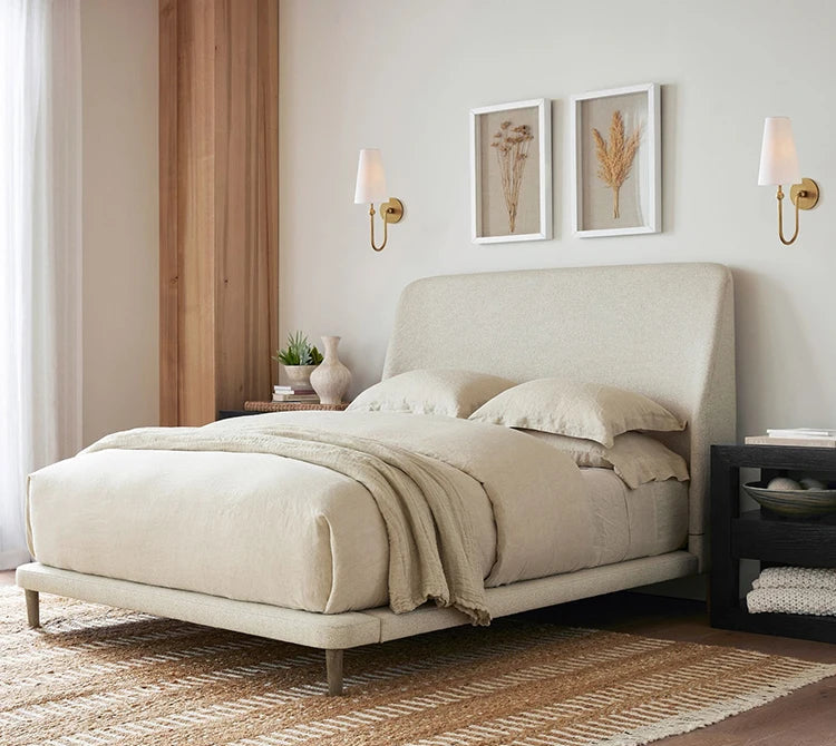 Beige upholstered bed in a bedroom setting with side tables and wall decor.