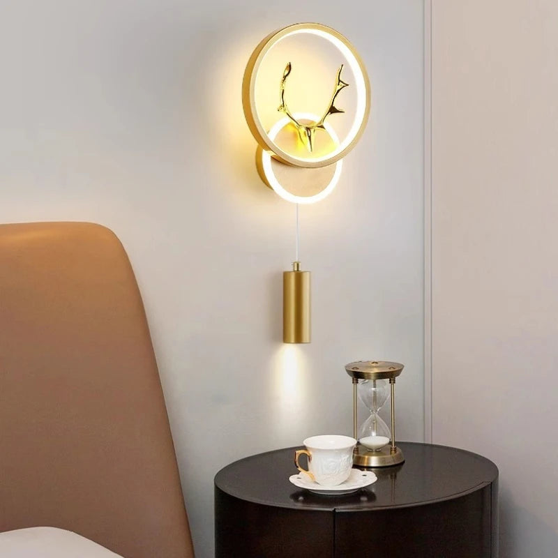 Gold wall sconce with circular LED halo and integrated antler design, mounted next to a bedside table with a teacup and hourglass.