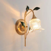 Gold wall sconce with a clear glass shade shaped like a flower and green leaf accents, mounted on a white wall with light casting upwards.