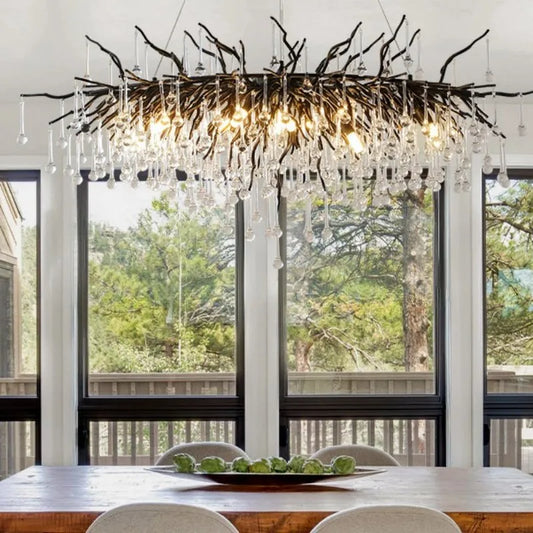 A modern black branch-style chandelier with numerous teardrop crystals hanging above a wooden dining table with a scenic forest view through large windows.