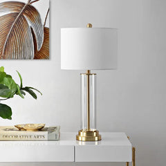 A clear glass cylinder table lamp with a brushed gold base and accents is displayed on a white surface with books and a plant.