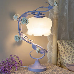 A purple table lamp with a floral rose vine design and a white frosted bell-shaped shade, illuminated on a bedside table.