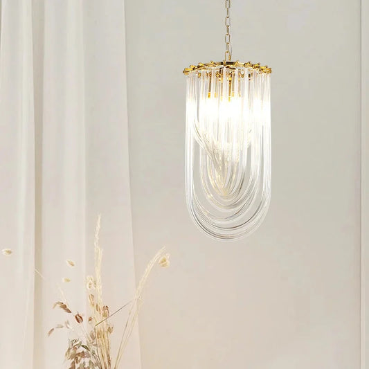 Close-up of a single gold U-shaped pendant light with clear glass tubes hanging from a chain against a white curtain background.