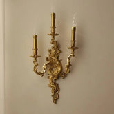 Gold candelabra-style wall sconce with three candle-shaped lights mounted on an ornate base against a light-colored wall.