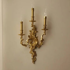 Gold candelabra-style wall sconce with three candle-shaped lights mounted on an ornate base against a light-colored wall.