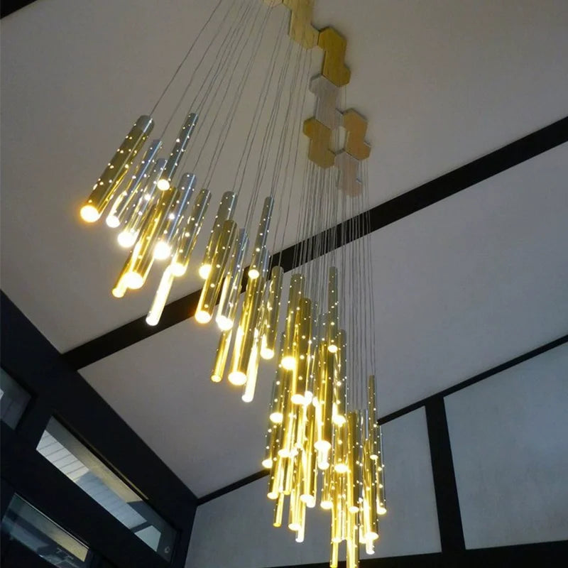 A low-angle shot looking up at a cascading chandelier with many thin, cylindrical tubes in gold and silver, illuminated and suspended from a geometric ceiling mount.