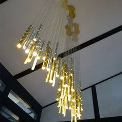 A low-angle shot looking up at a cascading chandelier with many thin, cylindrical tubes in gold and silver, illuminated and suspended from a geometric ceiling mount.
