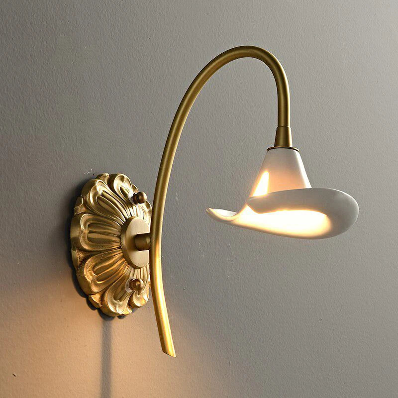 Close-up of a gold floral ceramic wall sconce, highlighting the intricate backplate and the smooth white ceramic shade emitting light.