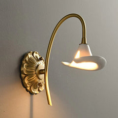 Close-up of a gold floral ceramic wall sconce, highlighting the intricate backplate and the smooth white ceramic shade emitting light.