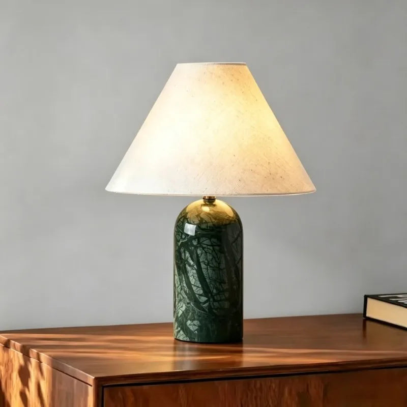 Green stone art ceramic table lamp with a fabric shade, illuminated and placed on a wooden cabinet.