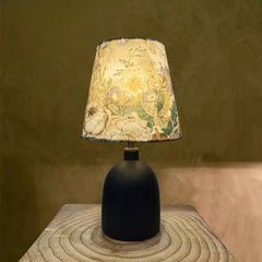 Close-up of a black ceramic table lamp with a floral fabric shade, illuminated in a warm setting against a textured wall.