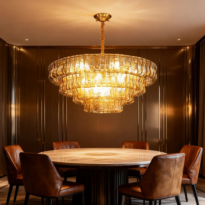 A multi-tiered gold chandelier with amber glass accents hanging over a round marble dining table with brown leather chairs.