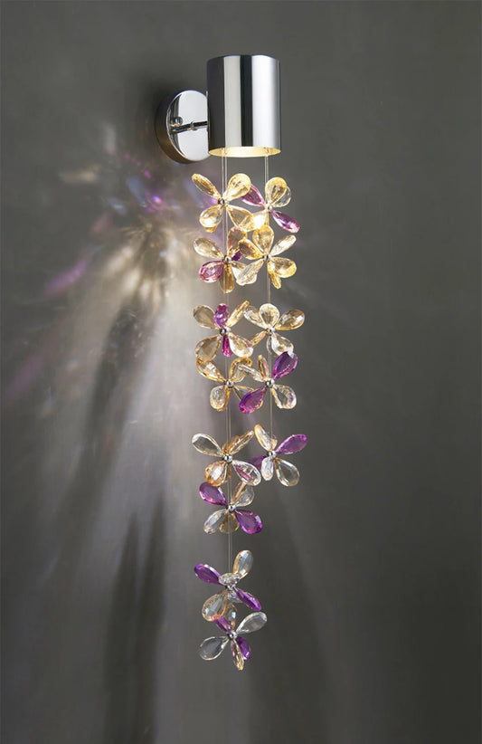 Polished chrome wall sconce with a cascading chain of clear and colored crystal flowers casting light patterns on a gray wall.