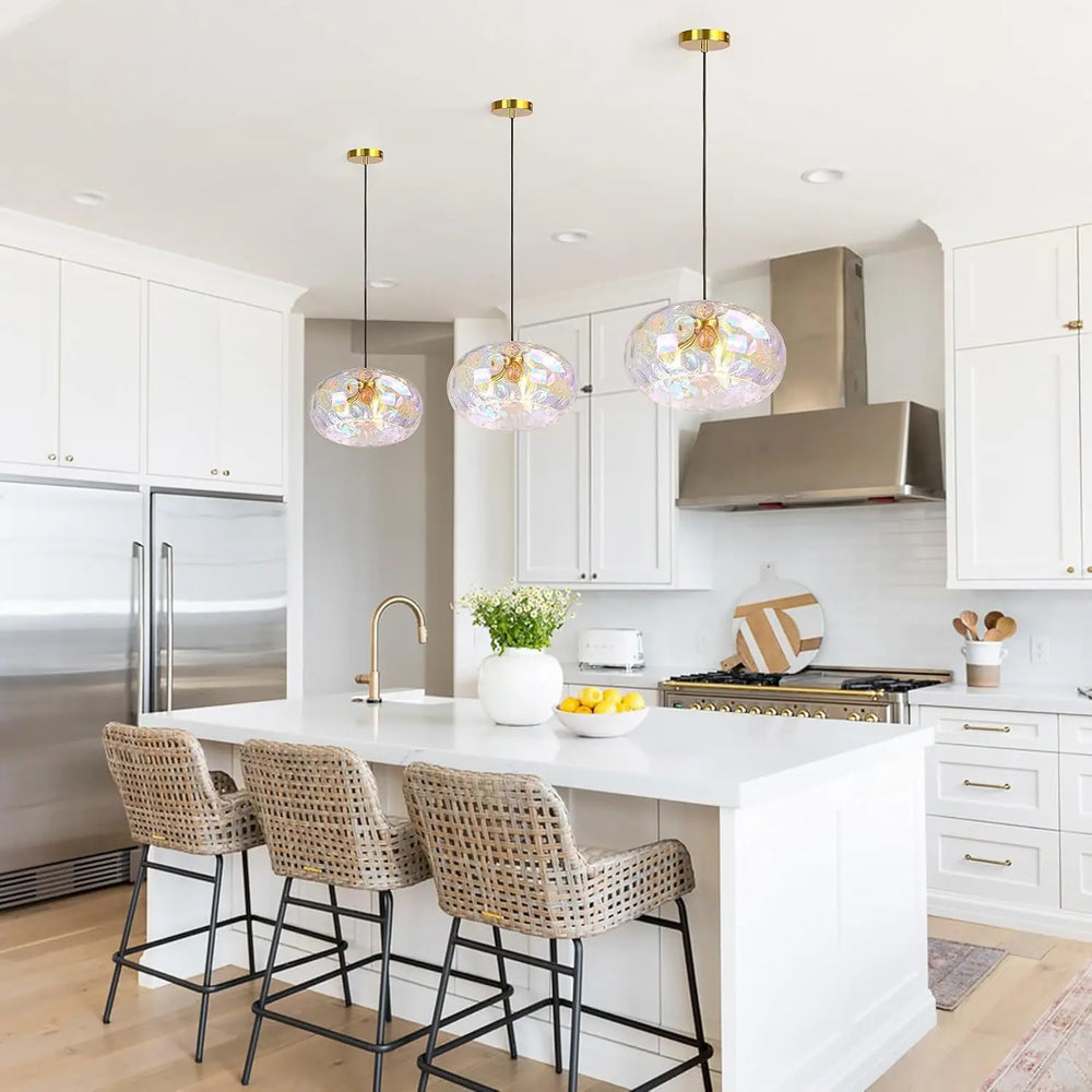 A bright white kitchen with a large island, three gold water ripple glass pendant lights hanging above it, and wicker bar stools.