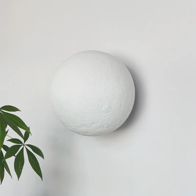 A white 3D printed moon shaped wall sconce with a textured surface is mounted on a white wall. A green leaf plant is in the lower left foreground.