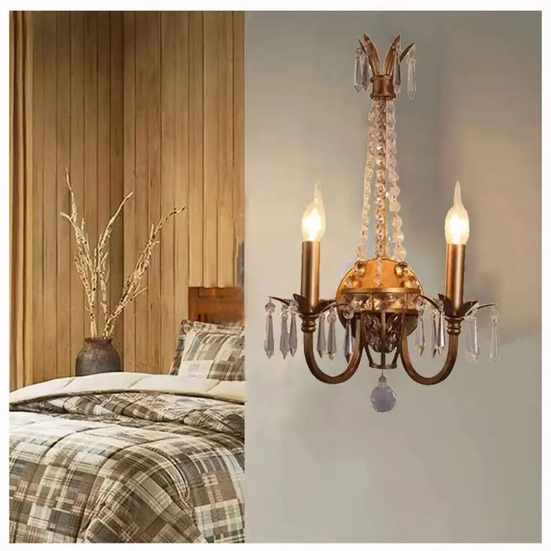 A gold crystal wall sconce with two candle-style lights mounted on a wall next to a bed with plaid bedding.