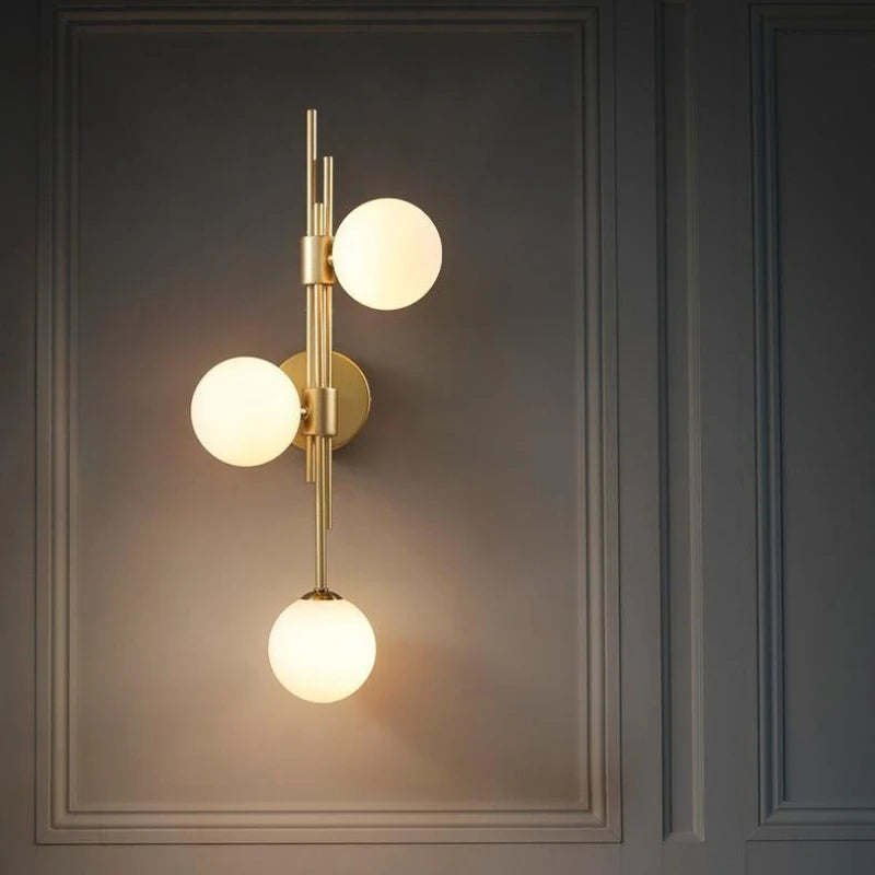 A modern gold wall sconce with three white frosted glass globes mounted vertically on a brass stem against a dark gray paneled wall.