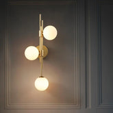 A modern gold wall sconce with three white frosted glass globes mounted vertically on a brass stem against a dark gray paneled wall.