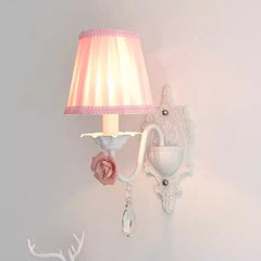 Close-up of a single white wall sconce with a pink pleated fabric shade, pink rose accent, and crystal droplet.