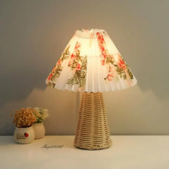 Table lamp with a floral print pleated fabric shade and a woven rattan base, lit on a table.