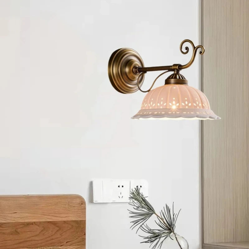 A gold and white ceramic floral wall sconce mounted on a white wall next to a power outlet and a plant.