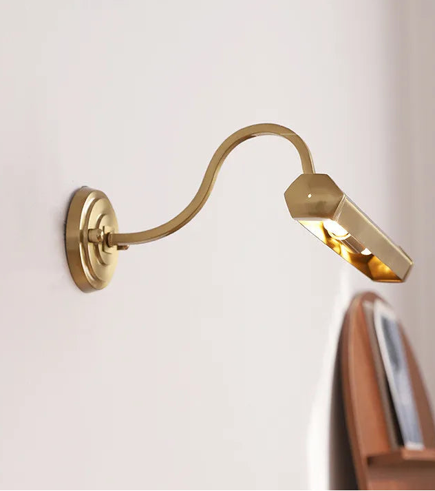 Gold wall-mounted lamp on a white wall with a wooden shelf in the background