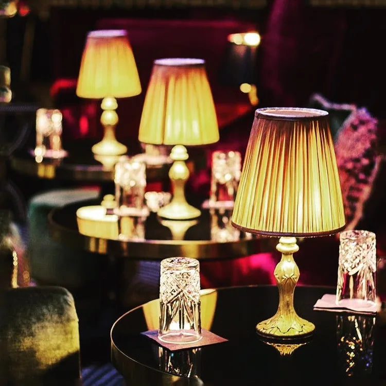 A dimly lit room with several round tables; each table has a lit gold-toned pleated fabric shade table lamp and glassware.