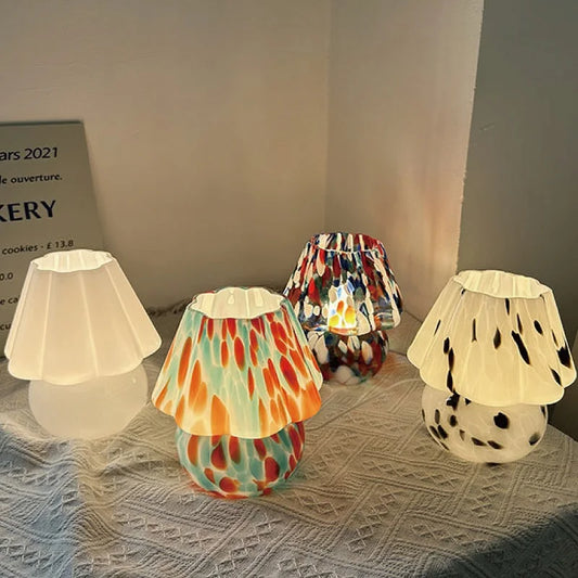 Four mushroom-shaped table lamps with stained glass shades in various colorful patterns and one with a white speckled pattern, arranged on a textured surface.