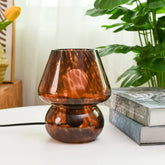 Brown glass table lamp on a white surface with books and a plant in the background