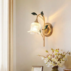Gold flower wall sconce with clear glass shade in a room setting with white walls, a cream curtain, and a vase of flowers on a table below.