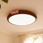 A round, flush mount ceiling light with a wood-tone frame and a pink bow accent, emitting a soft white light against a white ceiling.