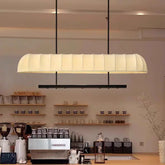 A long, rectangular, cream-colored fabric pendant light fixture with vertical ribbing, suspended by black cords over a cafe counter.