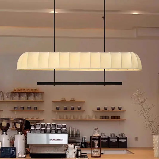 A long, rectangular, cream-colored fabric pendant light fixture with vertical ribbing, suspended by black cords over a cafe counter.