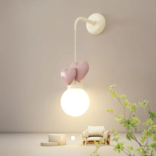 A wall sconce with a cream mounting bracket, a white cord, three pink heart-shaped accents, and a white globe light emitting warm light.