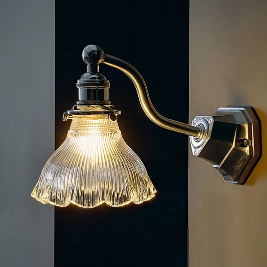 Close-up of a wall sconce with a clear, fluted glass shade and a curved chrome arm mounted on a dark wall.