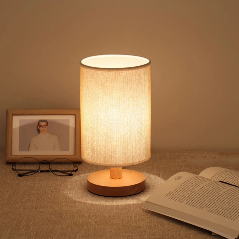 A linen drum shade table lamp with a plain fabric shade and a wooden base, placed on a textured surface next to an open book and eyeglasses.
