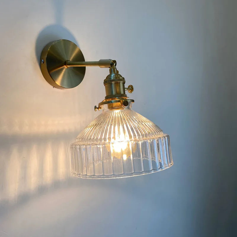 Clear fluted glass shade wall sconce with a brass arm and mounting plate illuminated against a beige wall, casting shadows.
