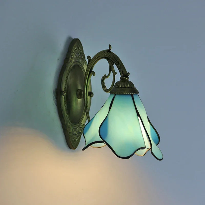 A light blue Tiffany-style wall sconce with a flower-shaped stained glass shade, showing the curve of the distressed gold metal arm against a gray wall.