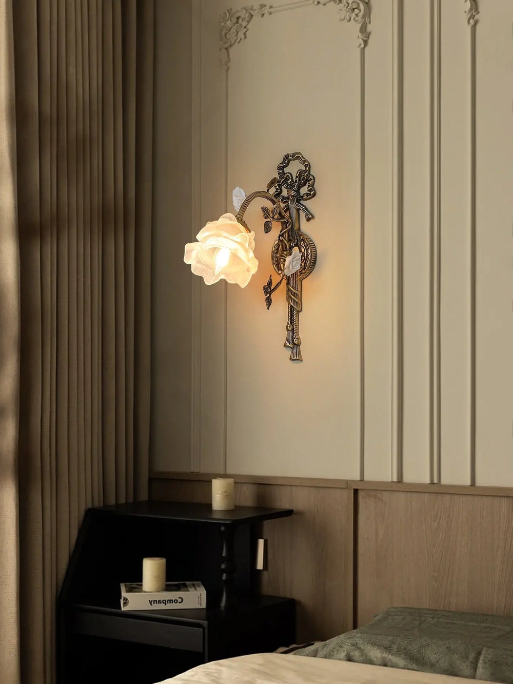 Close-up of an antique gold flower wall sconce with a white frosted glass shade, mounted on a wall with decorative paneling and a black nightstand beside it.