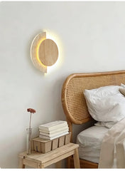 A modern wall sconce with a wood circle and speckled stone arc mounted on a wall above a bedside table with books.