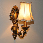 A single gold wall sconce with a cream fabric shade and ornate, antique-inspired detailing, mounted on a light-colored wall.