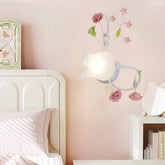 A white wall sconce with pink rose and green leaf accents, featuring a frosted glass shade, mounted on a pink wall next to a bed.