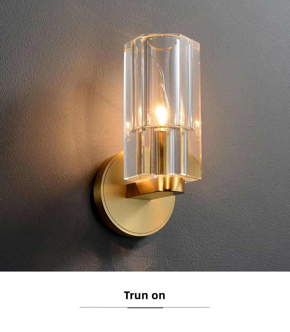 Wall-mounted light fixture with clear glass shade and gold metal base on a gray wall.
