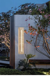 Rectangular wall sconce with a natural stone finish and a vertical LED light, mounted on a textured gray wall outdoors at dusk.